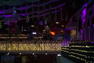 A festive indoor scene with a large Christmas tree adorned with lights and ornaments placed in the center. String lights illuminate the balcony area, casting a warm glow. Purple and pink lights enhance the ambiance, illuminating the arches of the building. Various decorations hang from the ceiling, adding to the holiday atmosphere.