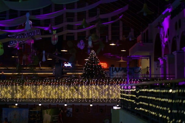 A festive indoor scene with a large Christmas tree adorned with lights and ornaments placed in the center. String lights illuminate the balcony area, casting a warm glow. Purple and pink lights enhance the ambiance, illuminating the arches of the building. Various decorations hang from the ceiling, adding to the holiday atmosphere.