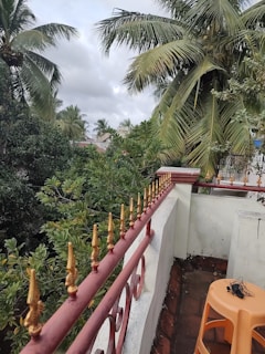 A small balcony bordered by ornate metal railings with gold-colored finials. Tall palm trees with lush green leaves are visible in the background under an overcast sky. A plastic stool, possibly orange in color, is placed on the balcony floor, with some wires or cords resting on it.