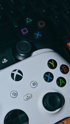 Two video game controllers are positioned close together. One is black with colorful geometric symbols, and the other is white with brightly colored buttons labeled X, Y, A, and B. They are resting on a surface that hints at a dark keyboard beneath.