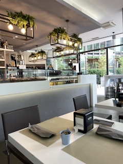 A modern, well-lit café interior with sleek furniture. The table settings include placemats, grey napkins, a napkin holder, and small decorative potted plants. The ceiling features stylish light fixtures with hanging greenery. Large windows allow natural light to illuminate the space, revealing a view of foliage outside. The counter holds a pastry display and assorted bottles.