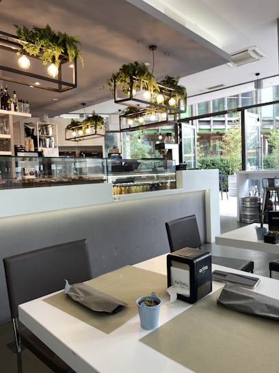 A modern, well-lit café interior with sleek furniture. The table settings include placemats, grey napkins, a napkin holder, and small decorative potted plants. The ceiling features stylish light fixtures with hanging greenery. Large windows allow natural light to illuminate the space, revealing a view of foliage outside. The counter holds a pastry display and assorted bottles.