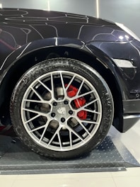 A close-up of a car wheel with a detailed and stylish alloy rim. The red brake calipers add a vivid contrast against the metallic finish of the rim. The car body appears sleek and shiny, with reflections visible, indicating a well-maintained or new vehicle. The floor under the wheel has a textured, metallic surface.