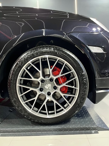 A close-up of a car wheel with a detailed and stylish alloy rim. The red brake calipers add a vivid contrast against the metallic finish of the rim. The car body appears sleek and shiny, with reflections visible, indicating a well-maintained or new vehicle. The floor under the wheel has a textured, metallic surface.