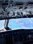 The image displays the cockpit of an aircraft with a detailed view of the control panels and flight instruments. A large and picturesque body of water is visible through the cockpit windows, with surrounding landforms.