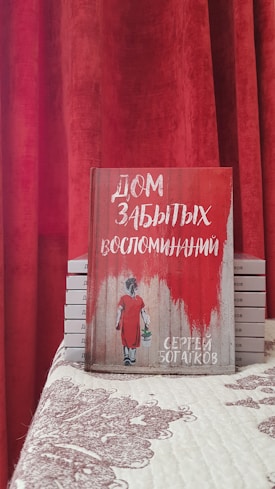 A book with a red and white cover is prominently displayed against a red curtain background. The book cover features an illustration of a person wearing a red coat and holding a green plant in a pot. The book is supported by a stack of other books with similar covers, and it is placed on a patterned tablecloth.