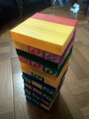 A tower of colorful blocks arranged neatly, each block displaying a number. The blocks are stacked in alternating colors of red, yellow, green, and blue. The background includes a wooden floor and some indistinct items to the side.