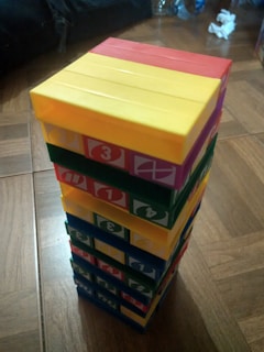 A tower of colorful blocks arranged neatly, each block displaying a number. The blocks are stacked in alternating colors of red, yellow, green, and blue. The background includes a wooden floor and some indistinct items to the side.