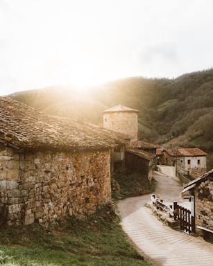 A quaint village scene featuring stone buildings with tiled roofs set against a backdrop of rolling hills. The sun is setting or rising, casting a warm glow across the landscape and highlighting the rustic texture of the stonework. A winding path leads through the village, bordered by grass and more stone structures.
