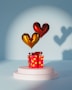 A red gift box with yellow polka dots sits on a white pedestal. Above the box are two heart-shaped balloons, one gold and one red, casting distinct shadows on a blue background.