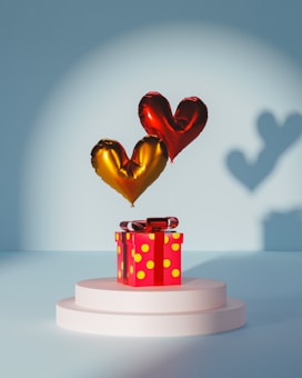 A red gift box with yellow polka dots sits on a white pedestal. Above the box are two heart-shaped balloons, one gold and one red, casting distinct shadows on a blue background.