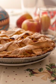A freshly baked apple pie with a golden-brown lattice crust sits on a table. The pie is placed on a festive, holiday-themed tablecloth, which features holly and ribbon patterns. In the background, there are a few whole apples and a glass, slightly out of focus, adding to the cozy and inviting atmosphere.