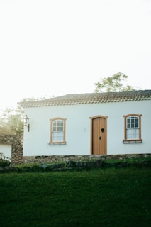 A charming, small house with white walls and orange-brown window and door frames is situated on a grassy area. The house features two windows with decorative grids and a wooden door. A stone wall and street lamp add rustic elements, while trees and foliage are visible in the background, contributing to a tranquil setting.
