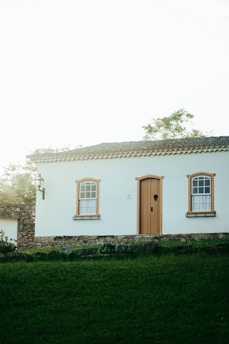 A charming, small house with white walls and orange-brown window and door frames is situated on a grassy area. The house features two windows with decorative grids and a wooden door. A stone wall and street lamp add rustic elements, while trees and foliage are visible in the background, contributing to a tranquil setting.