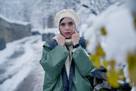 A person stands in a snowy outdoor setting wearing a warm green coat and cream-colored knit accessories, including a hat and scarf. Snowflakes fall around the individual, who stands near a stone wall partially covered in snow. A blurred background suggests a wintry landscape with trees covered in snow.