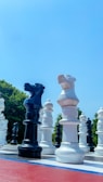 A large outdoor chessboard with oversized black and white chess pieces is displayed. The pieces are standing on a checkered surface with a clear blue sky overhead. Trees are visible in the background.