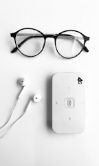 A pair of round black-rimmed eyeglasses, a pair of white earphones, and a white portable Wi-Fi router labeled '4G' are neatly arranged on a plain white background.