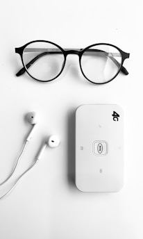 A pair of round black-rimmed eyeglasses, a pair of white earphones, and a white portable Wi-Fi router labeled '4G' are neatly arranged on a plain white background.