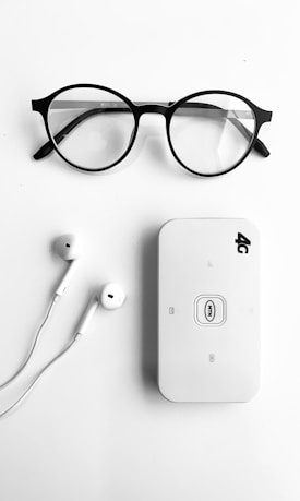 A pair of round black-rimmed eyeglasses, a pair of white earphones, and a white portable Wi-Fi router labeled '4G' are neatly arranged on a plain white background.