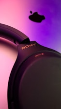 A close-up view of a set of black Sony headphones against a vibrant gradient background of pink and purple shades. Above the headphones, a black Apple logo is visible, creating a contrast with the bright background.