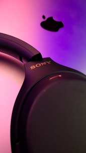 A close-up view of a set of black Sony headphones against a vibrant gradient background of pink and purple shades. Above the headphones, a black Apple logo is visible, creating a contrast with the bright background.