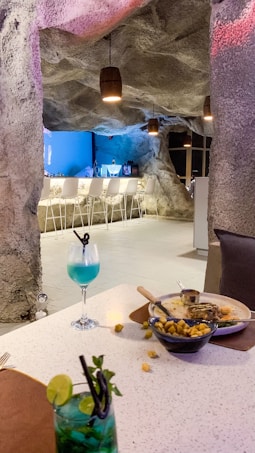 A cave-themed bar with a rock-textured interior, featuring a modern setup with a long white counter surrounded by high white chairs. Hanging lantern-style lights add warm lighting. On the table in the foreground, there are two colorful cocktails and a partially eaten meal, including a skillet and some scattered fried pieces.