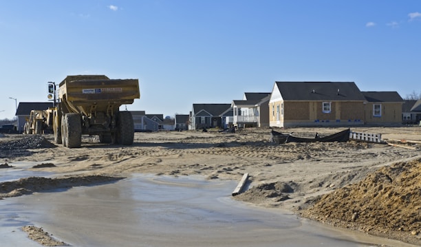 A construction site with partially built houses in a residential area. The foreground features a large yellow dump truck on sandy and muddy terrain. Several houses under construction are visible in the background, with some appearing more complete than others. The sky is clear and blue, indicating good weather.