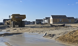 A construction site with partially built houses in a residential area. The foreground features a large yellow dump truck on sandy and muddy terrain. Several houses under construction are visible in the background, with some appearing more complete than others. The sky is clear and blue, indicating good weather.