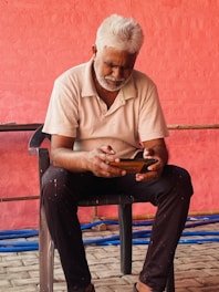 An older man with white hair and a beard is seated on a black chair, focused on using a mobile phone. He is dressed in a white polo shirt and dark pants. The background features a textured, red-painted wall with some blue pipes on the ground.