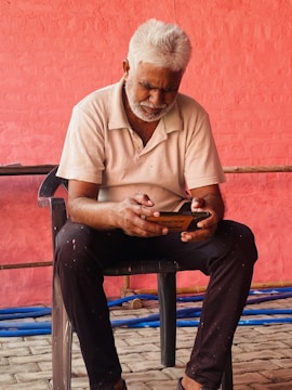 An older man with white hair and a beard is seated on a black chair, focused on using a mobile phone. He is dressed in a white polo shirt and dark pants. The background features a textured, red-painted wall with some blue pipes on the ground.