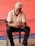 An older man with white hair and a beard is seated on a black chair, focused on using a mobile phone. He is dressed in a white polo shirt and dark pants. The background features a textured, red-painted wall with some blue pipes on the ground.