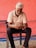 An older man with white hair and a beard is seated on a black chair, focused on using a mobile phone. He is dressed in a white polo shirt and dark pants. The background features a textured, red-painted wall with some blue pipes on the ground.