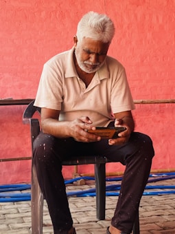 An older man with white hair and a beard is seated on a black chair, focused on using a mobile phone. He is dressed in a white polo shirt and dark pants. The background features a textured, red-painted wall with some blue pipes on the ground.