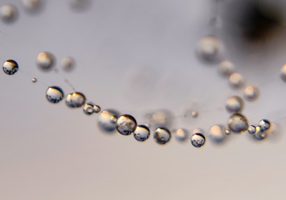 A close-up view of several water droplets delicately suspended on a spider web, with each droplet acting as a tiny lens reflecting its surroundings. The background is softly blurred, enhancing the focus on the clarity and spherical shapes of the droplets.