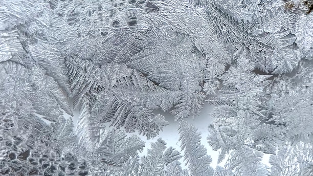Intricate patterns of frost crystals form on a surface, resembling delicate ferns or leaves. The crystalline structures are detailed and finely textured, with overlapping and interlocking forms that create a natural lace-like effect.
