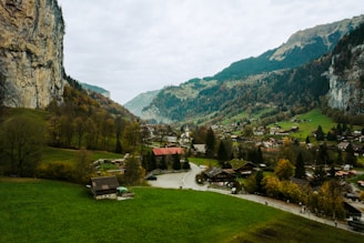 A scenic rural landscape featuring lush green fields, a village with scattered houses, and a winding road. The scene is bordered by majestic cliffs on the left and mountains in the background, partially covered with forests. The village appears peaceful and tranquil, surrounded by natural beauty.