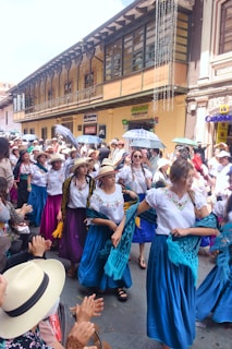 A vibrant street parade featuring women in colorful traditional outfits and hats. They are dancing and holding scarves, surrounded by an applauding crowd. The architecture in the background includes a yellow building with large windows and festive decorations.