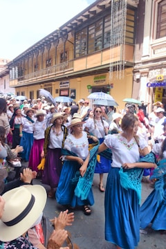 A vibrant street parade featuring women in colorful traditional outfits and hats. They are dancing and holding scarves, surrounded by an applauding crowd. The architecture in the background includes a yellow building with large windows and festive decorations.