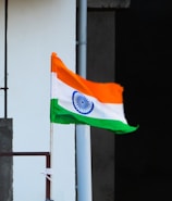 A flag featuring orange, white, and green horizontal stripes with a navy blue wheel in the center of the white stripe is mounted on a pole. The flag is waving in the wind against a concrete wall backdrop.