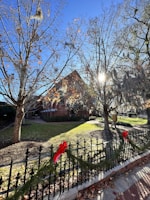 A scenic view of a residential area featuring a brick house partially obscured by leafless trees. Sunlight peeks through the branches, creating a bright and serene atmosphere. The fence in the foreground is decorated with festive garlands and red bows, hinting at a holiday season. The ground is covered with a mix of grass and fallen leaves.