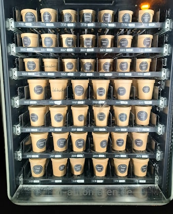 A vending machine filled with rows of brown paper cups, each labeled with different flavors and prices. The cups are neatly organized within clear slots across several rows and columns.
