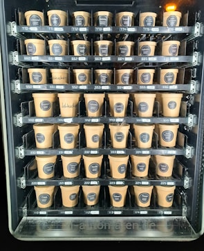 A vending machine filled with rows of brown paper cups, each labeled with different flavors and prices. The cups are neatly organized within clear slots across several rows and columns.