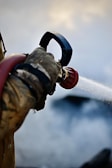 A close-up view of a gloved hand holding a hose nozzle that is spraying water, with water jets clearly visible against a blurred background.