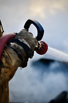 A close-up view of a gloved hand holding a hose nozzle that is spraying water, with water jets clearly visible against a blurred background.
