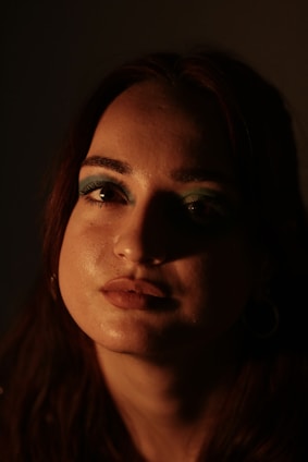 A close-up portrait of a person with intense lighting that creates dramatic shadows on their face. The makeup is vibrant, with bright blue eyeshadow highlighting their eyes. The lighting emphasizes one half of the face, casting the other half in shadow, creating a mysterious and introspective atmosphere.