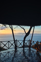An open pavilion with a tiled floor overlooks a peaceful ocean view at sunset. The sky is a blend of soft pastel colors with hues of orange, pink, and blue. A tree is visible through the wooden railing, adding a natural element to the scene.