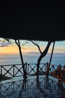 An open pavilion with a tiled floor overlooks a peaceful ocean view at sunset. The sky is a blend of soft pastel colors with hues of orange, pink, and blue. A tree is visible through the wooden railing, adding a natural element to the scene.