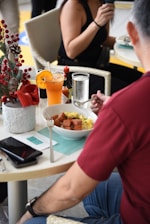 A person wearing a red shirt is seated at a table with a bowl of food, possibly containing scrambled eggs and diced meat. An orange drink with a slice of orange, a bottle of Tabasco sauce, and a glass of water are on the table. A white potted plant with red decorations is nearby. Another person in a black top is in the background holding a beverage.