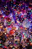 A vibrant display of holiday decorations with numerous colorful lights and ornaments hanging on tree branches. The scene is filled with sparkling bokeh effects, creating a festive and cheerful atmosphere.