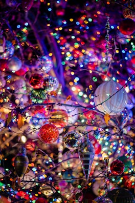 A vibrant display of holiday decorations with numerous colorful lights and ornaments hanging on tree branches. The scene is filled with sparkling bokeh effects, creating a festive and cheerful atmosphere.